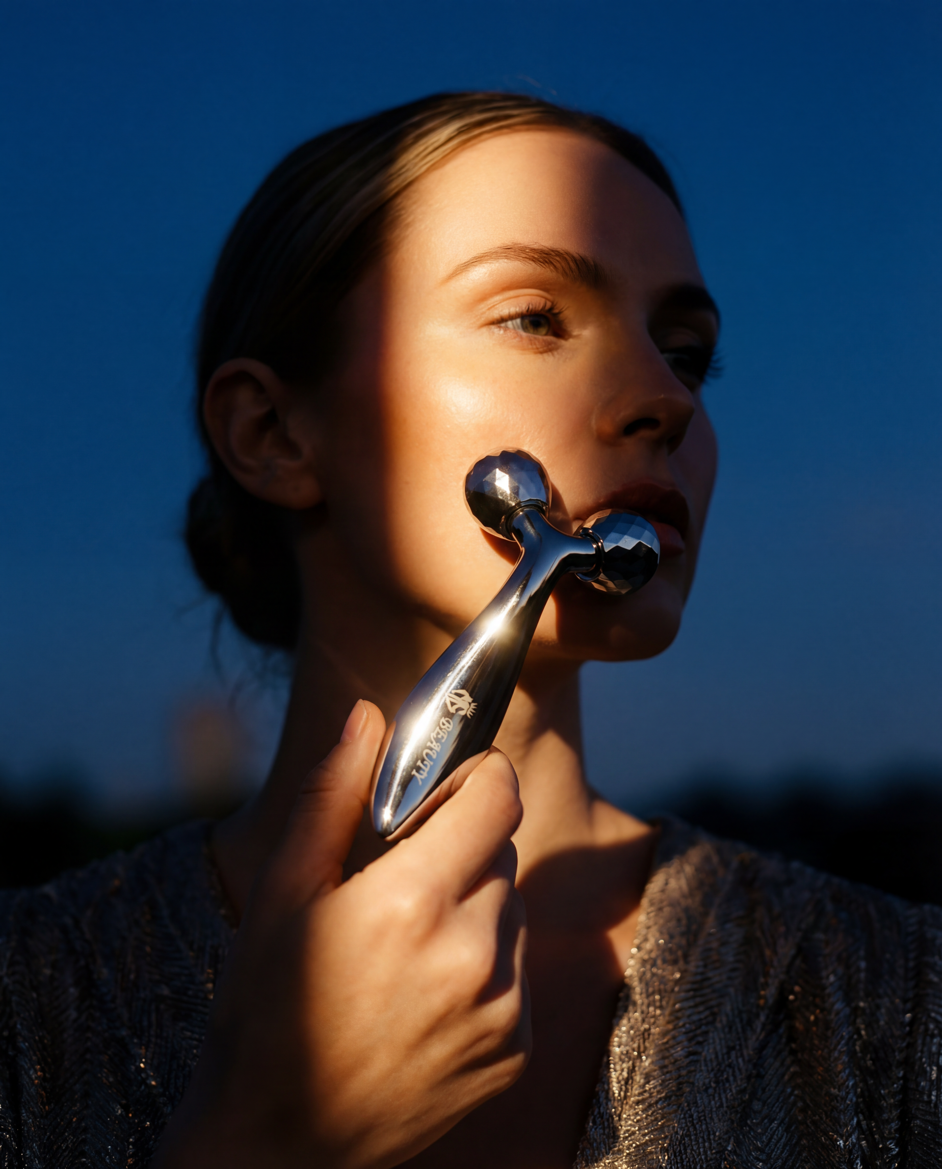 Woman holding a silver facial roller against her face with a dark blue background